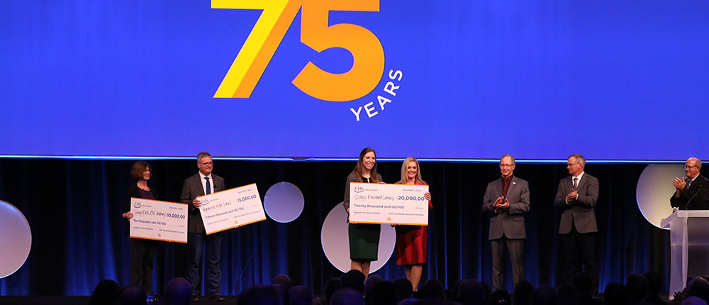 People in business attire standing on a stage holding oversized checks, the sign behind them reads 75 years