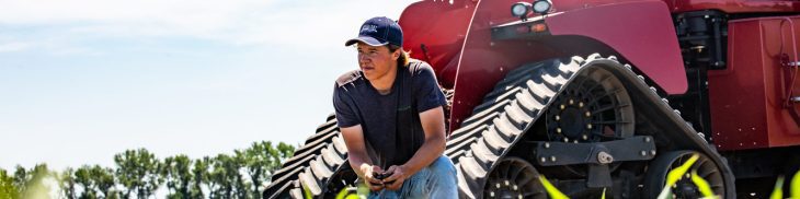 A teen kneels in front of a tractor and looks off into the distance